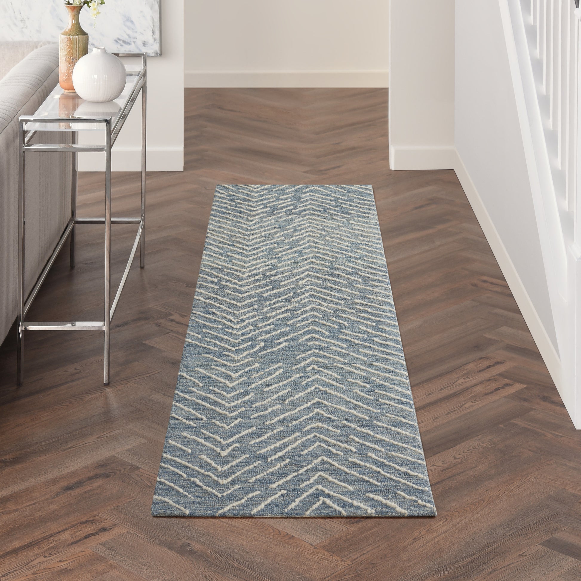 Blue chevron-pattern hallway runner rug on dark wood herringbone floor beside a metal console table with vases.