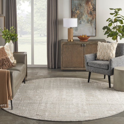 Round beige patterned area rug in a modern neutral living room with grey armchair, taupe sofa, wooden cabinet, and plants.