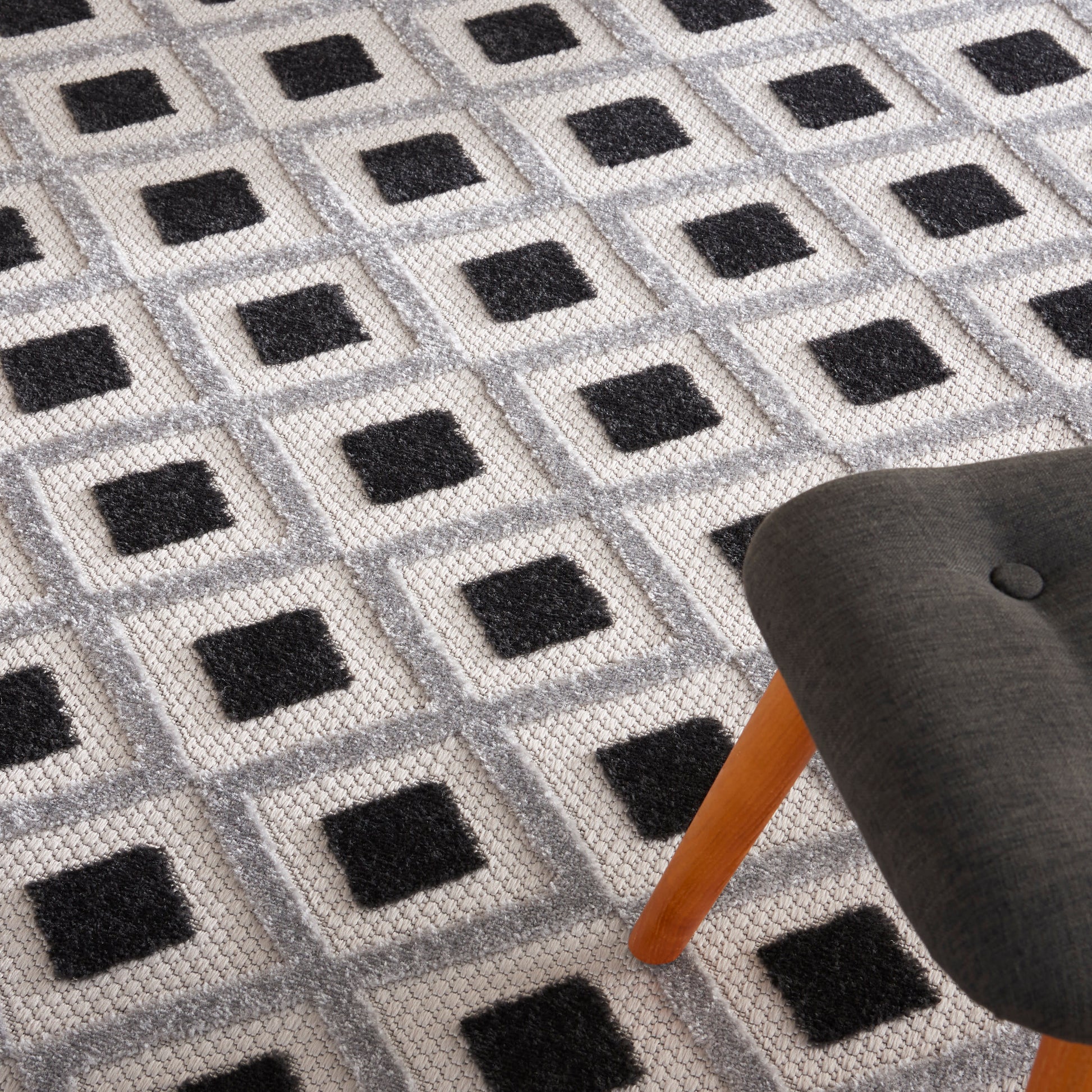 Geometric rug with light gray base and dark charcoal square motifs in a grid; plush pile beside a gray upholstered chair.