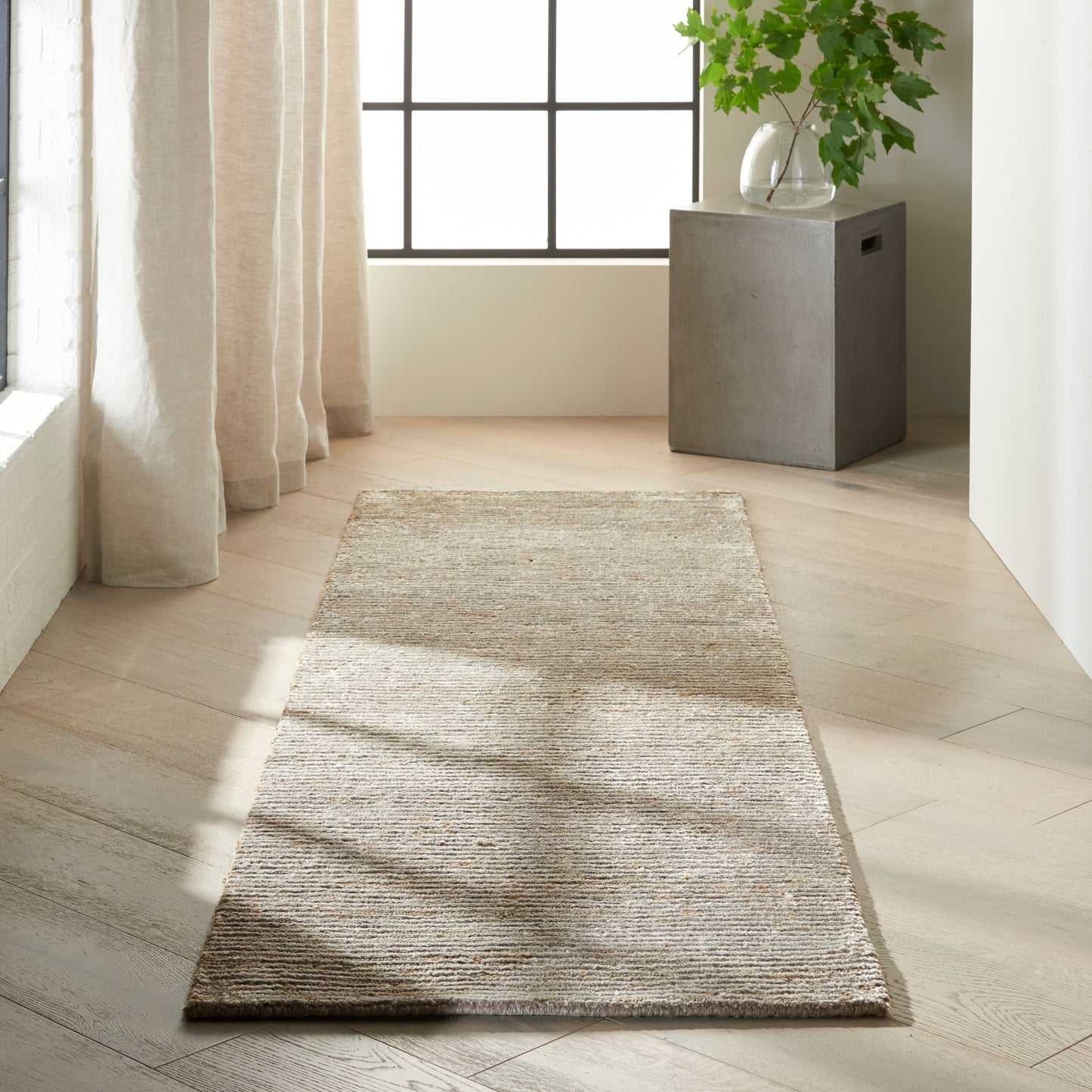 Beige woven runner rug on light wood floor in minimalist entryway with a grid window, curtains, and a concrete plant stand.