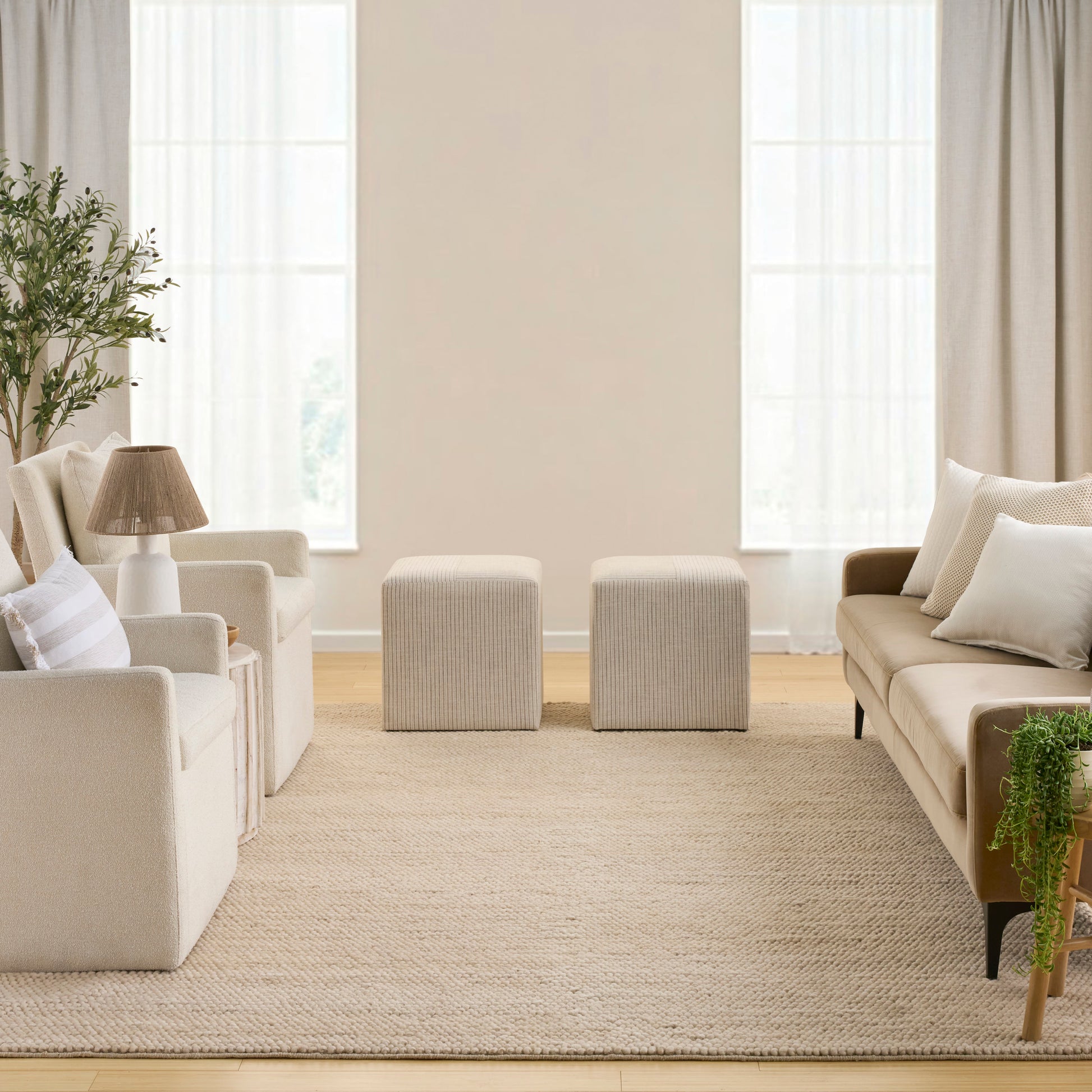 Neutral living room with cream sofas, two square beige ottomans, woven rug, potted plant, and natural light.