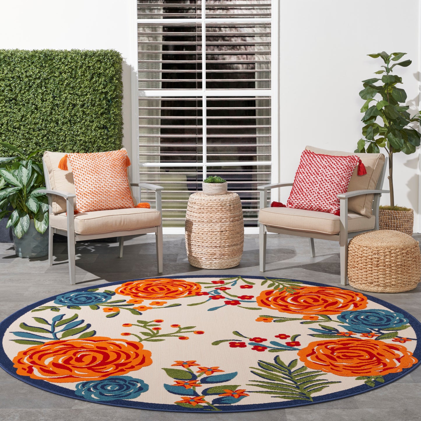 Outdoor patio with beige armchairs, orange & red cushions, a round floral rug, woven side tables, & greenery.