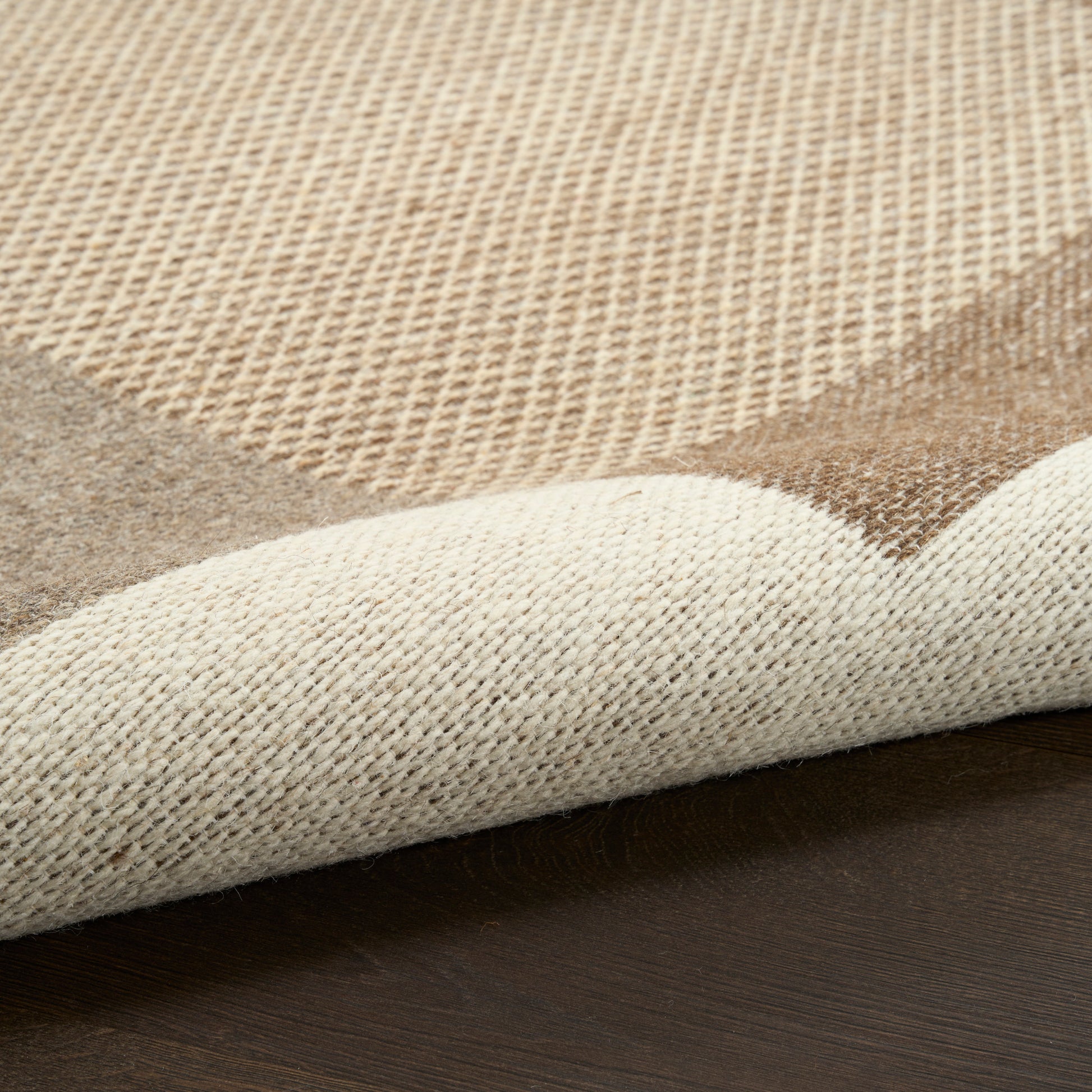 Beige and brown woven rug close-up with a rolled edge on dark wood floor; textured fibers.