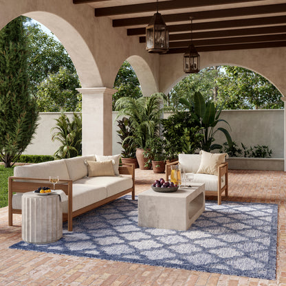 Patio lounge set with beige cushioned sofas, concrete coffee table, blue patterned rug, and potted plants under arches.