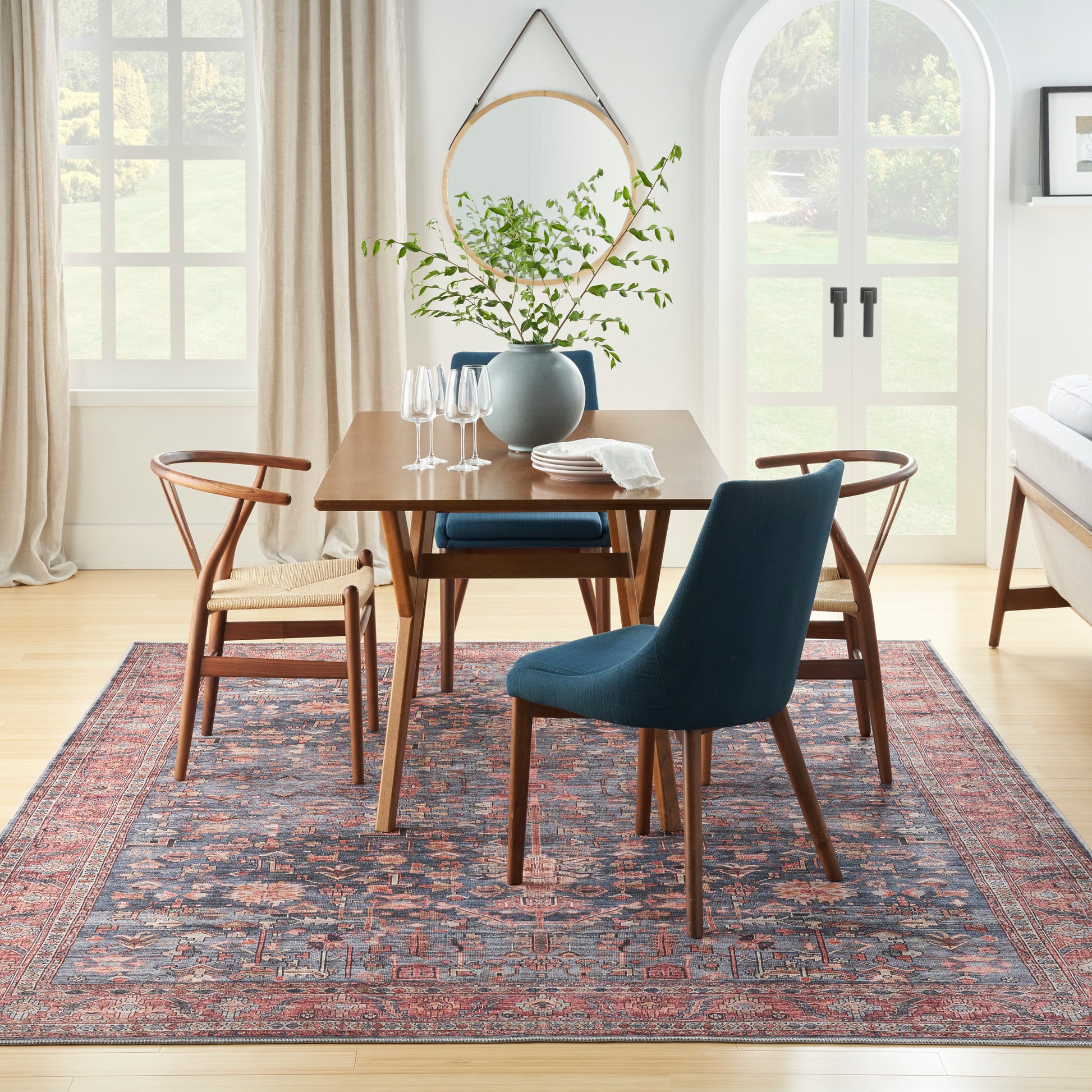 Wood dining table with blue upholstered chair and wooden spindle chairs on a Persian rug; greenery vase centerpiece.