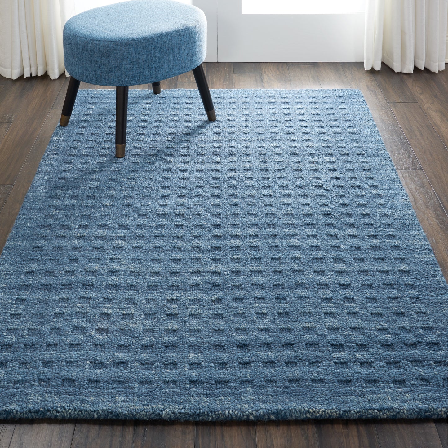 Blue textured rug with raised square pattern on dark wood floor, paired with a round blue ottoman.