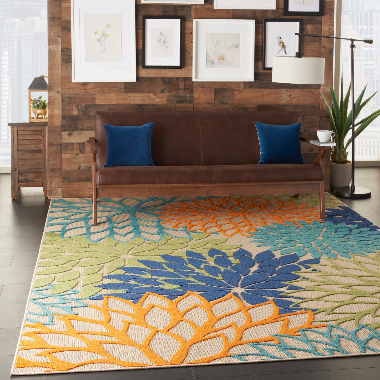 Large woven area rug with teal, orange, blue and lime tropical leaf motifs on a light background under a brown sofa.