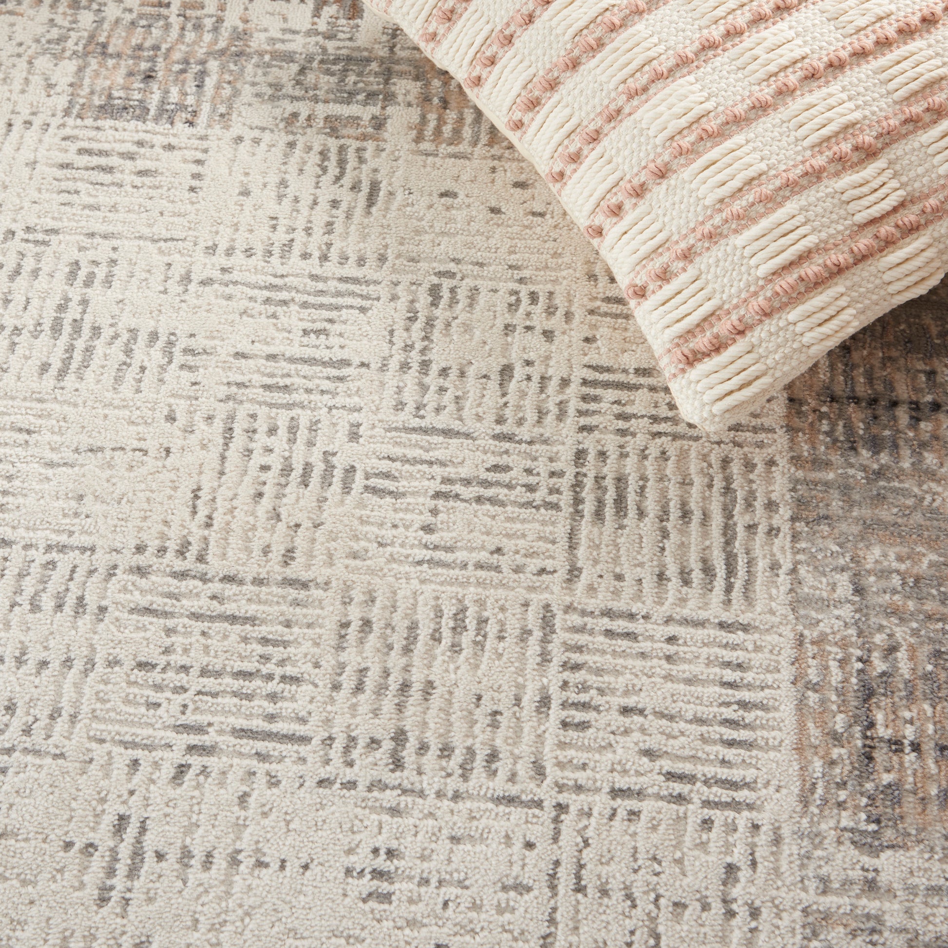 Beige-gray geometric area rug with square linear pattern; a pink striped knit blanket draped at the top corner.