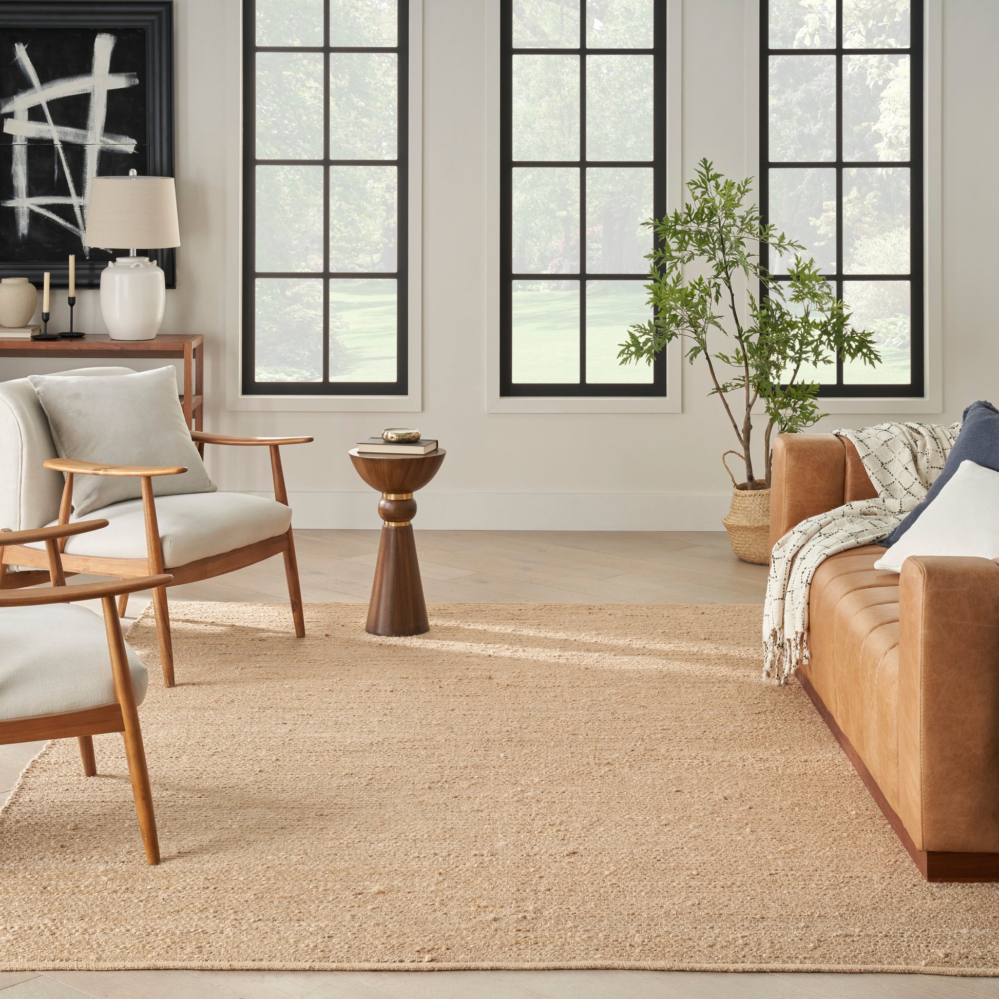 Bright living room with wooden chairs, white cushions, tan leather sofa, jute rug, black-framed windows and plants.
