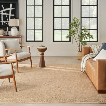 Bright living room with wooden chairs, white cushions, tan leather sofa, jute rug, black-framed windows and plants.