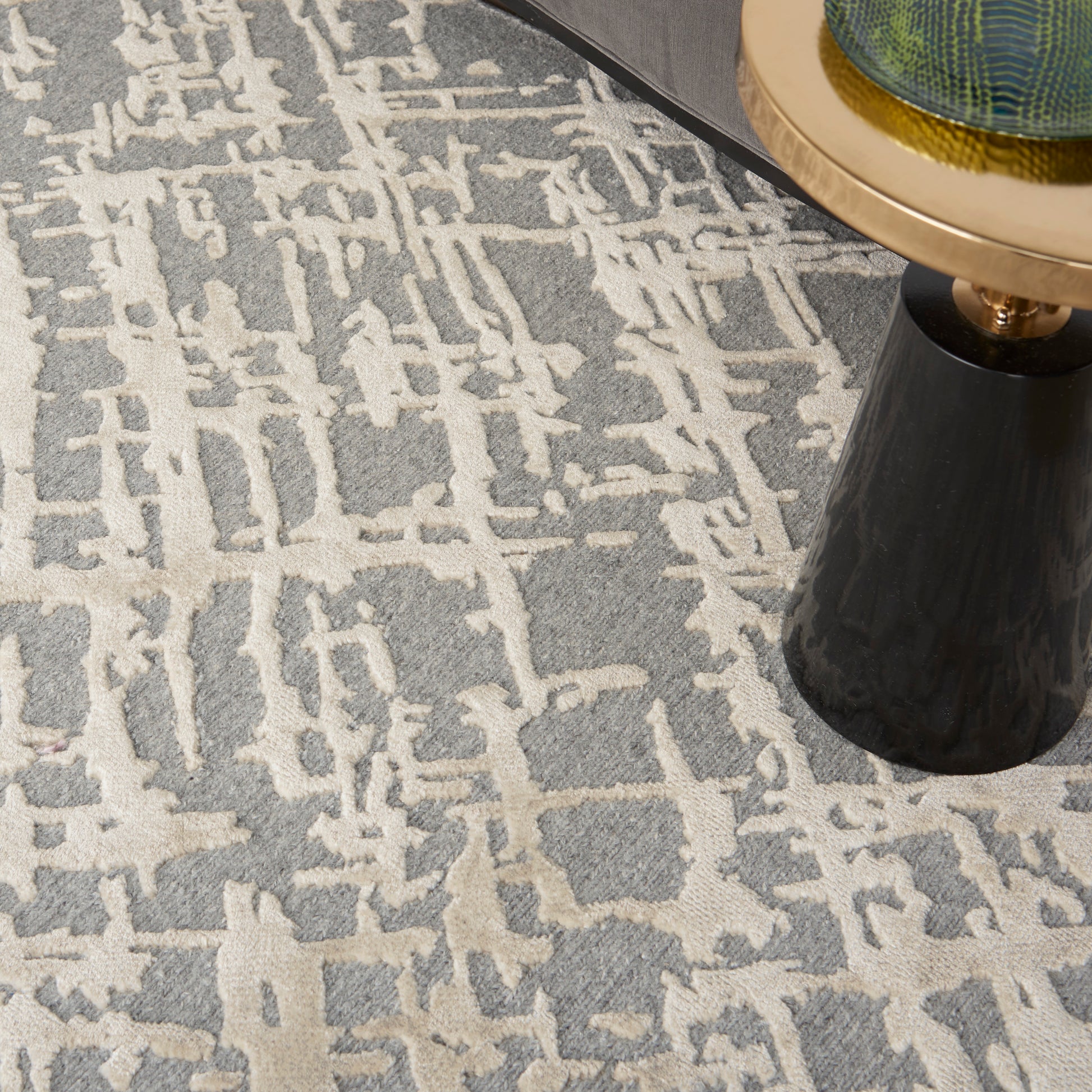 Gray rug with beige abstract geometric pattern; black tapered table leg with brass ring and green glass top.