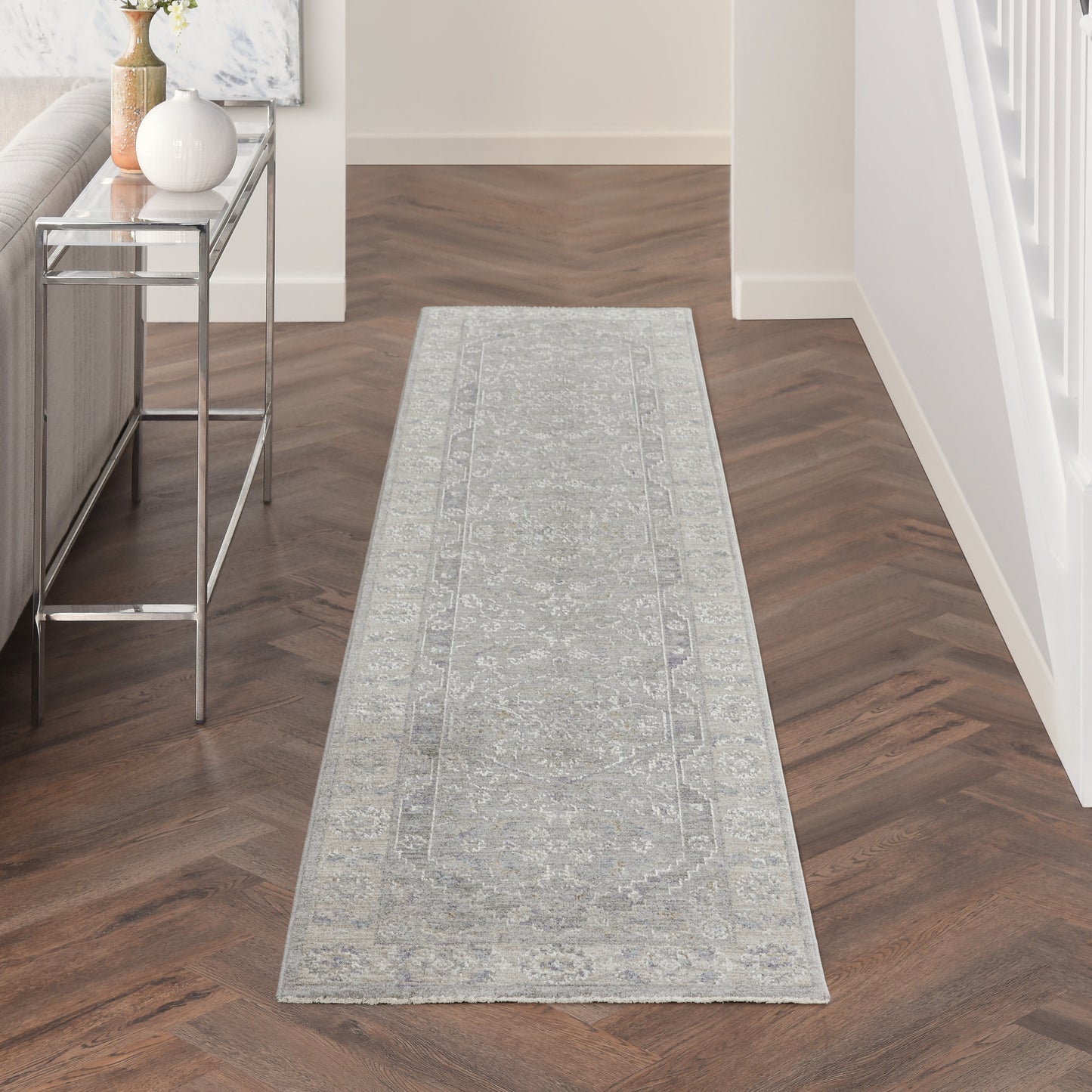 Light gray runner rug with subtle oriental pattern on herringbone wood hallway floor; metal console table with vase left.