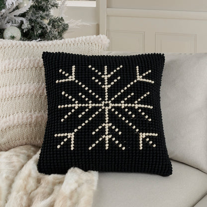 Black knitted square throw pillow with white snowflake motif; cozy holiday decor on light sofa with cream throw.