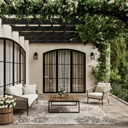 Coastal-style rug on an outdoor patio with beige cushioned wicker seating, wooden coffee table, arched black-framed doors & a green leafy pergola.