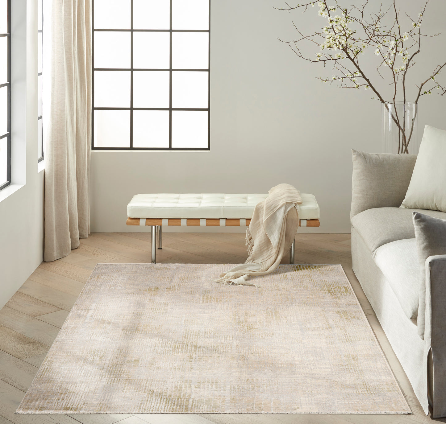 Neutral living room with large grid window, cream tufted bench, beige rug, light wood floors, gray sofa and plant branches.