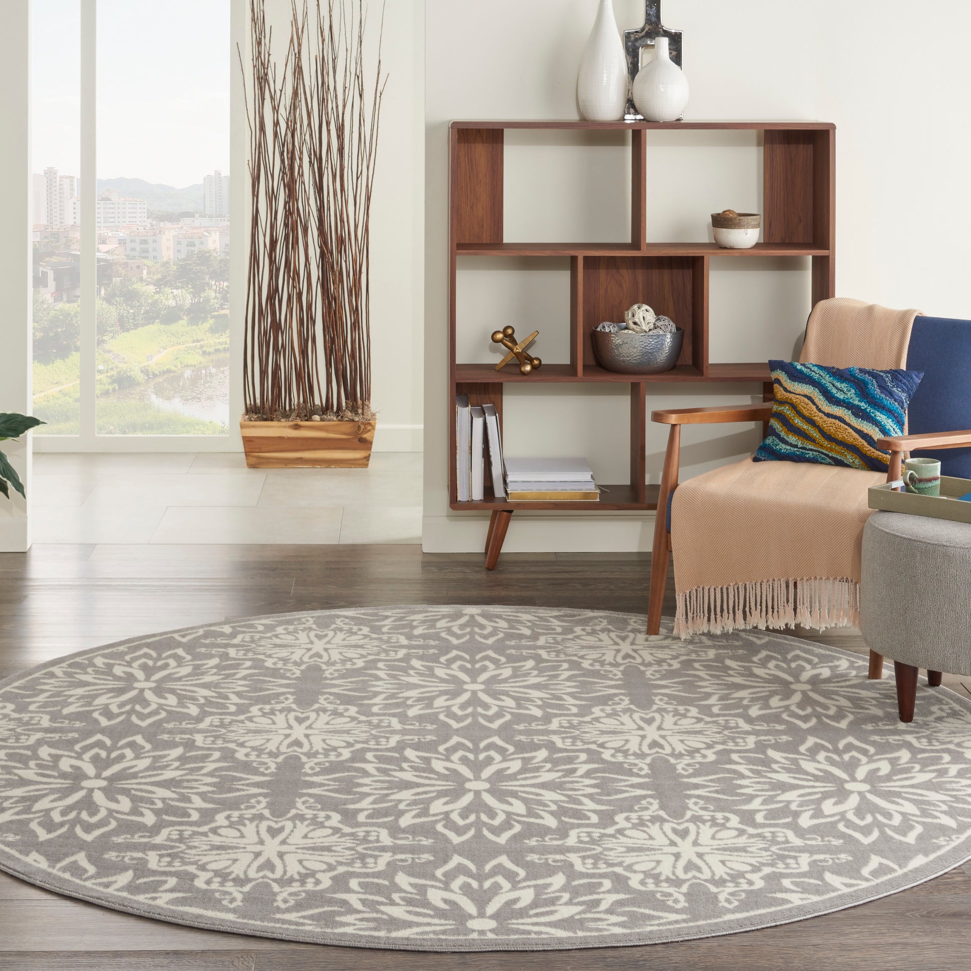 Round gray patterned rug in a modern living room with a wood bookshelf, beige throw, and twig planter.