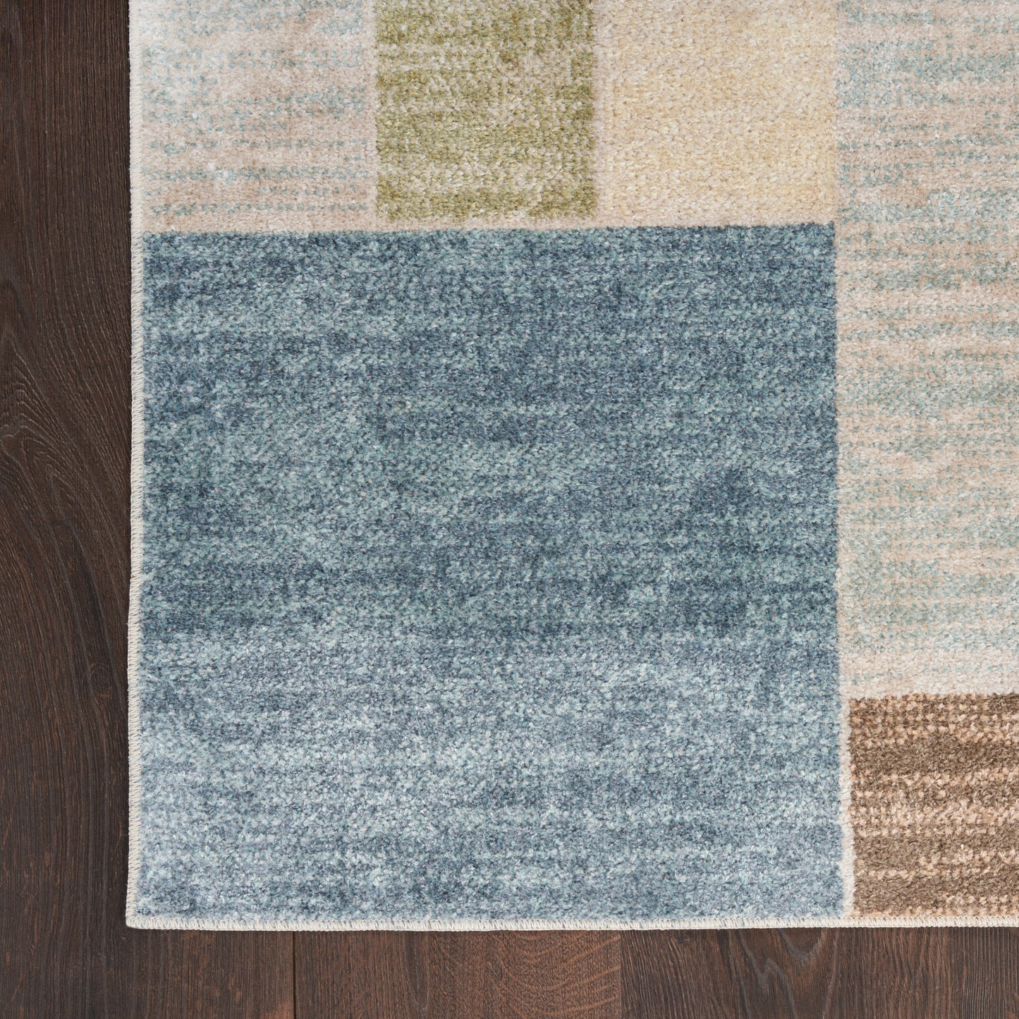Geometric patchwork rug with a blue center square and pastel blocks in beige, tan, green and light blue on dark wood floor.
