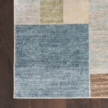 Geometric patchwork rug with a blue center square and pastel blocks in beige, tan, green and light blue on dark wood floor.