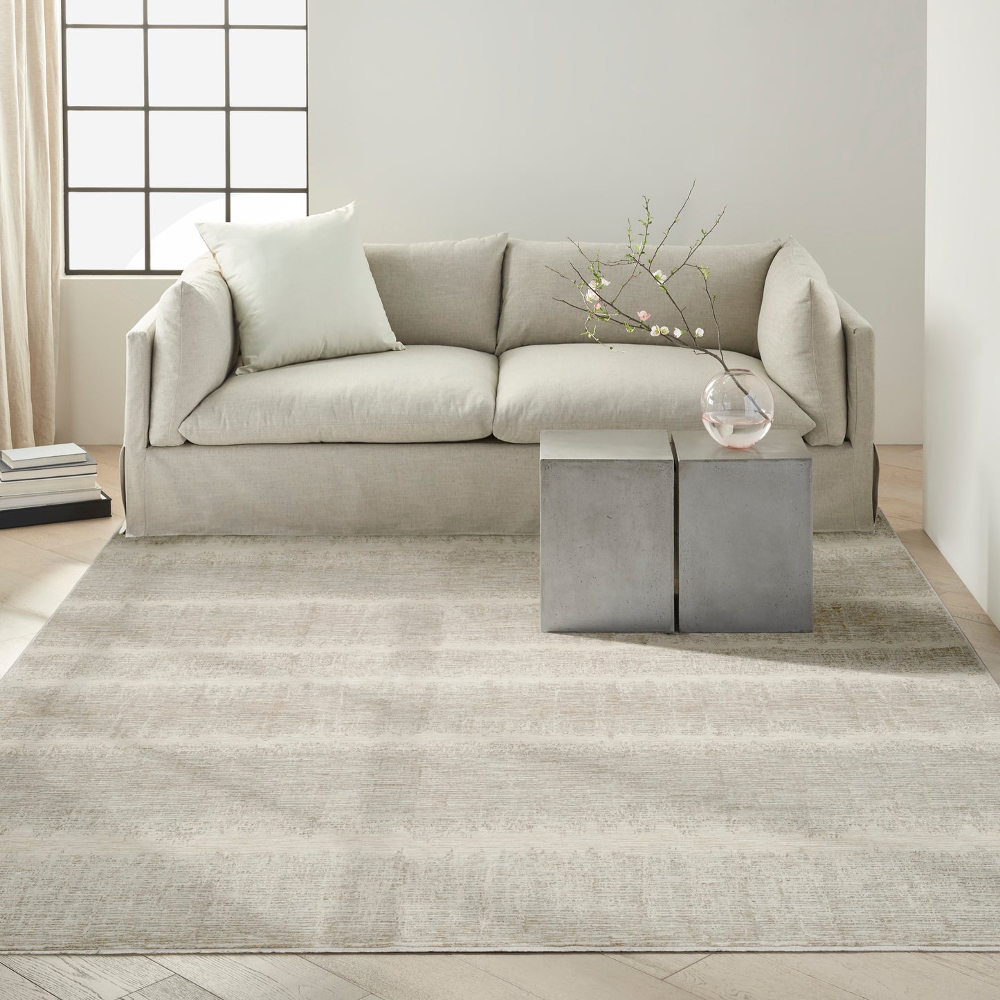 Beige fabric sofa with pale cushion, neutral rug, square concrete-like side tables, glass vase with flowering branches.
