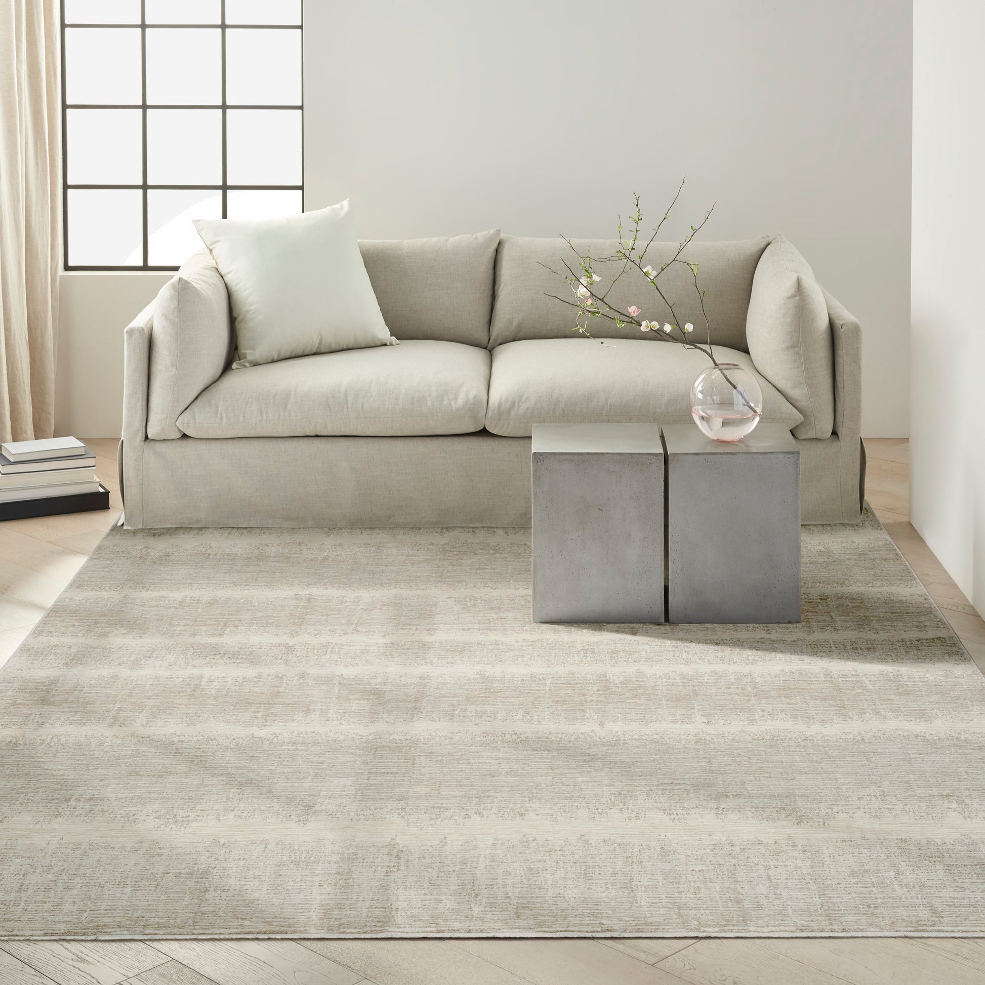Beige fabric sofa with pale cushion, neutral rug, square concrete-like side tables, glass vase with flowering branches.
