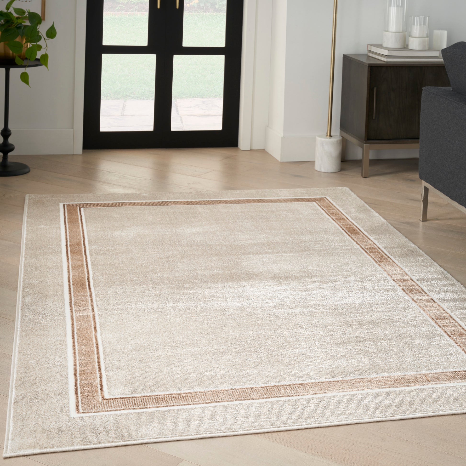 Rectangular beige area rug with a dual taupe border in a modern living room with light wood floor.