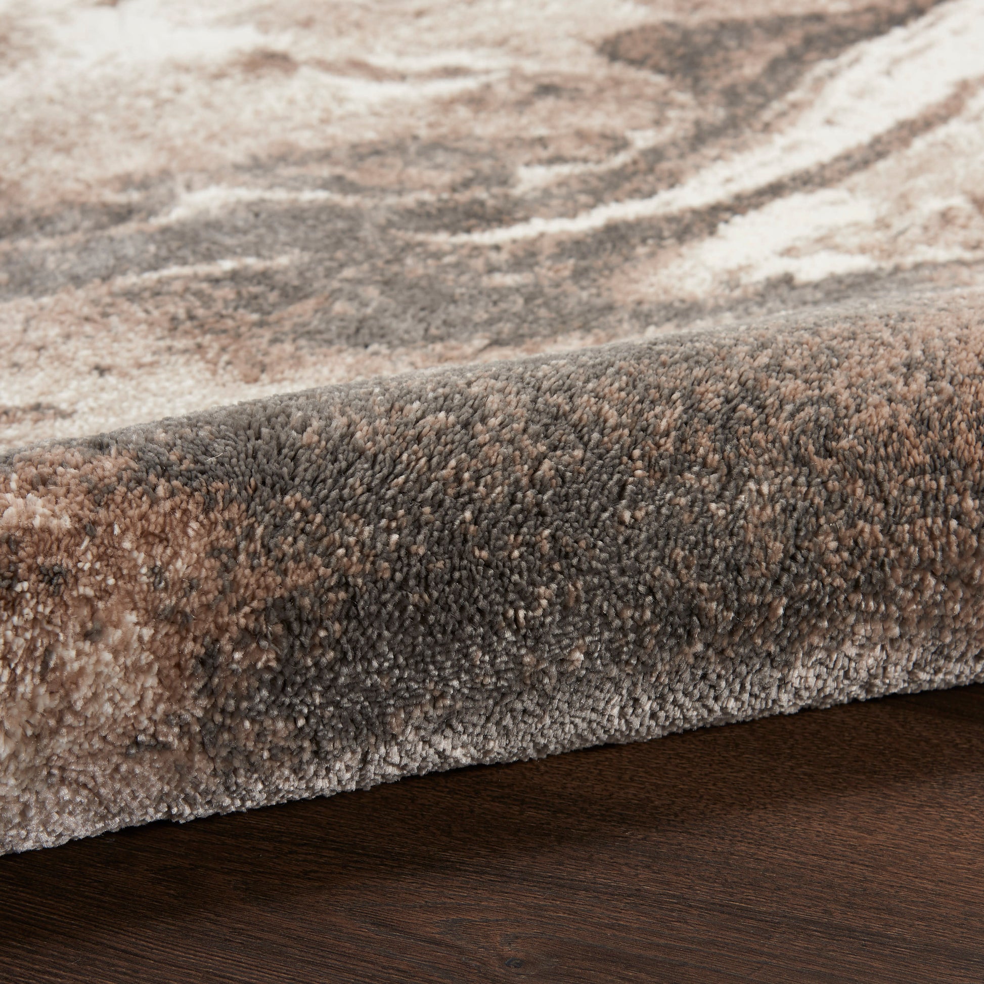 Close-up of a plush area rug with beige, brown & taupe tones, folded edge on dark wood floor.