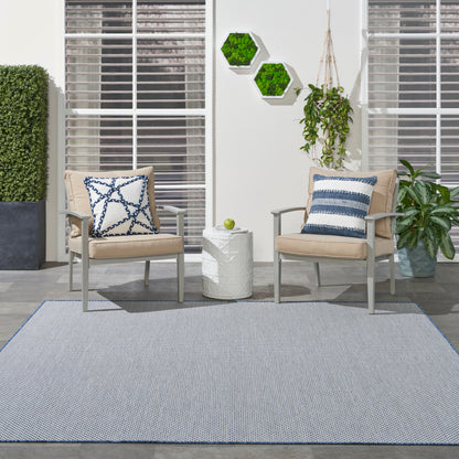 Modern outdoor seating with beige cushioned metal chairs, blue-white pillows, white side table, gray rug with blue border.
