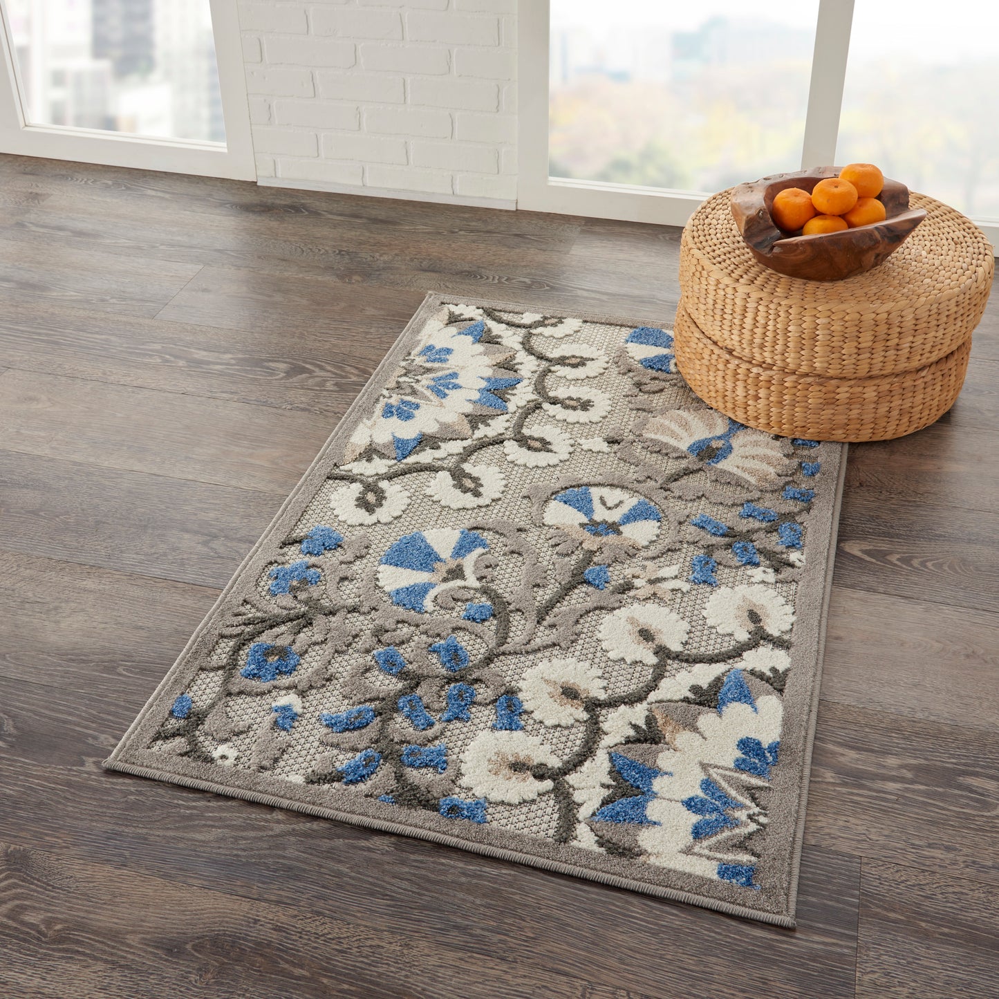 Rectangular floral area rug with gray background, cream and blue flowers, on dark wood floor beside a round woven basket with oranges.