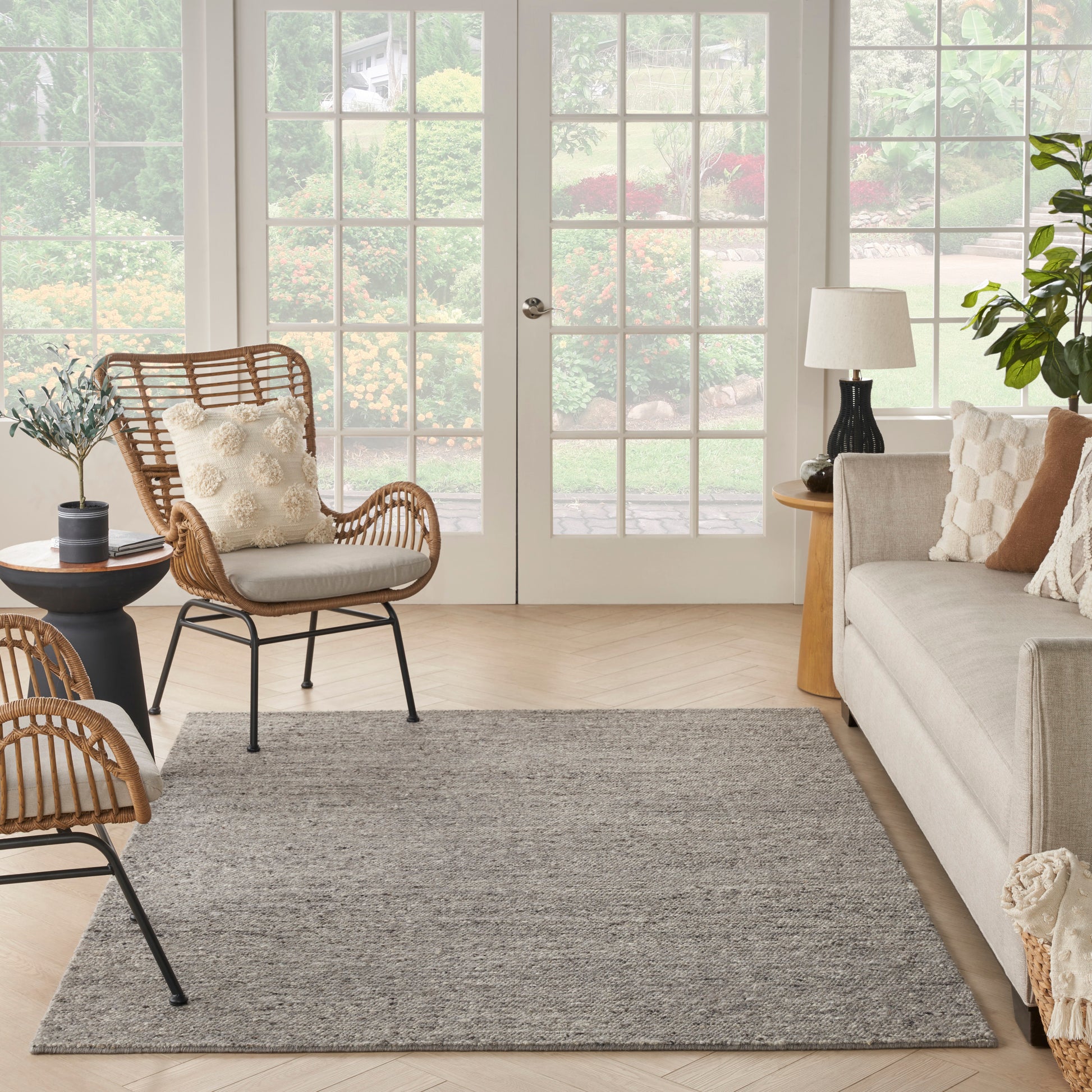 Bright living room with beige sofa, rattan chairs, gray rug, glass French doors to garden, greenery, and warm wooden accents.