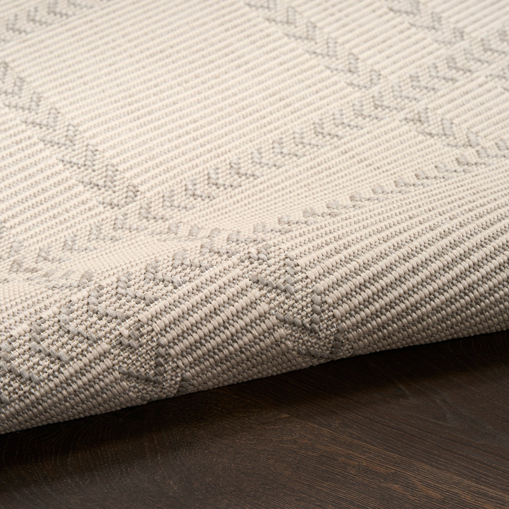 Beige woven area rug with subtle geometric trellis pattern, rolled edge on dark wood floor.