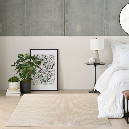 Neutral bedroom corner with white bedding, black-framed abstract art, potted plant, round black side table with lamp, beige rug.