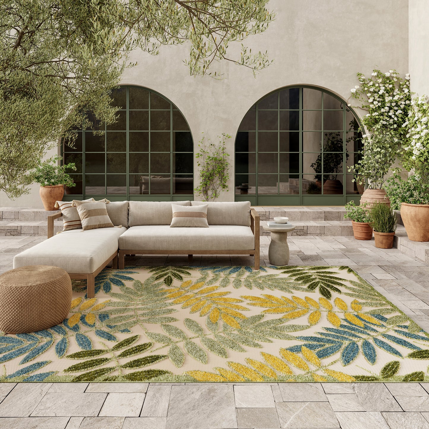 Outdoor seating area with beige sectional sofa on a leaf-pattern rug in green, yellow and blue; arched doors and potted plants.