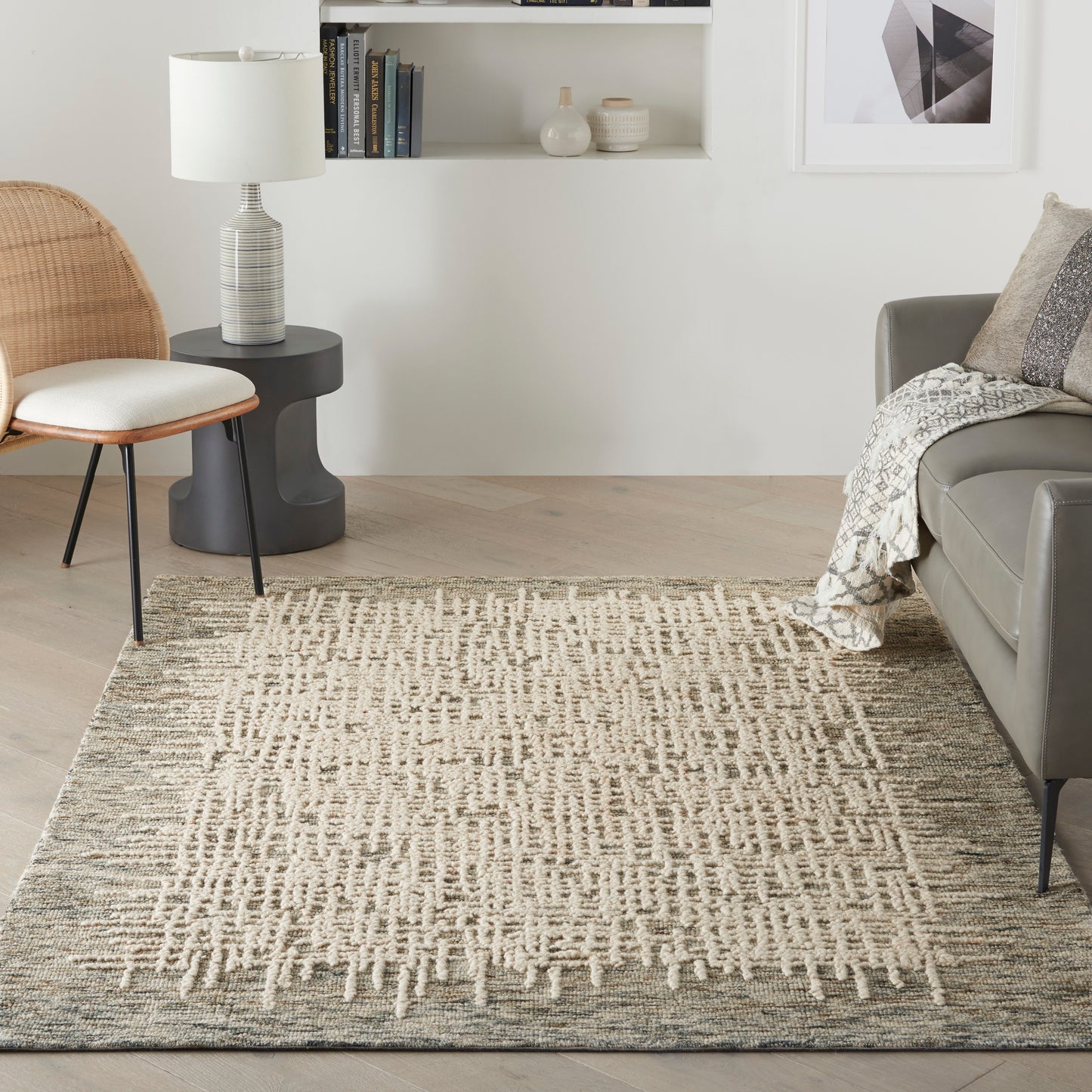 Neutral living room with a textured beige geometric rug, gray sofa, rattan chair, black side table, lamp, and white wall shelves.