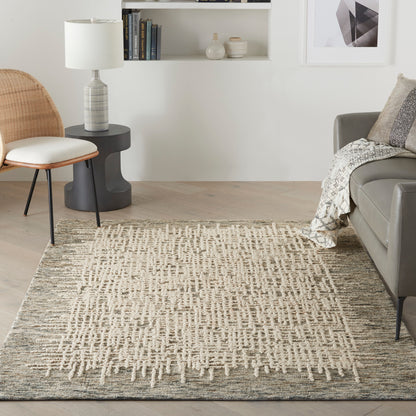 Neutral living room with a textured beige geometric rug, gray sofa, rattan chair, black side table, lamp, and white wall shelves.