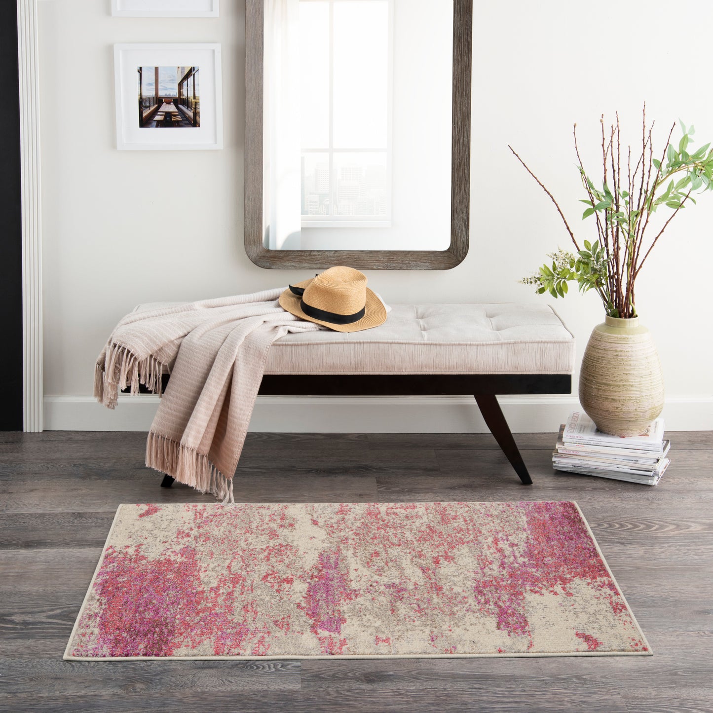 Beige tufted bench with dark wood legs, draped blanket and hat; pink-beige abstract rug; vase with branches, wall mirror.