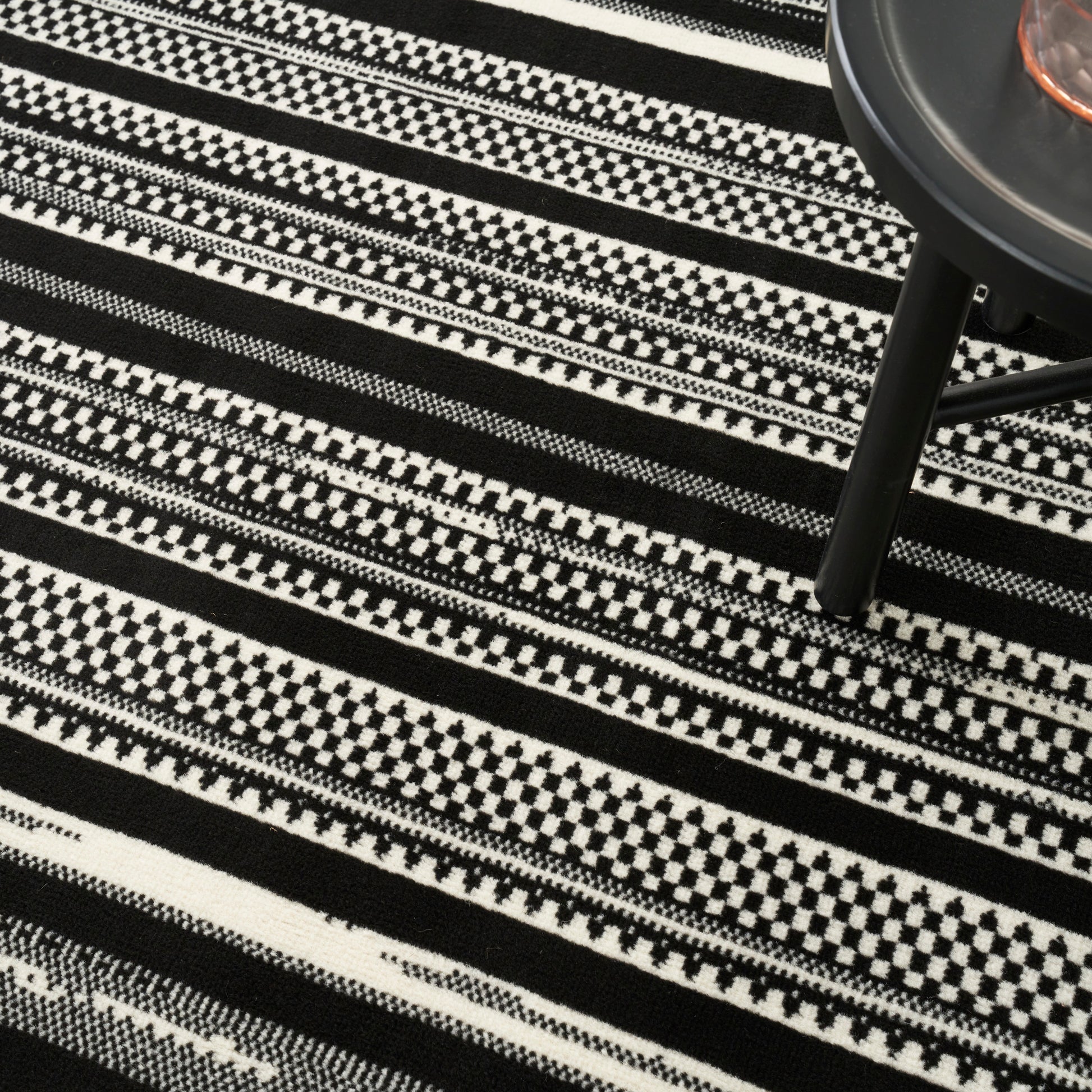 Black and white geometric striped woven rug with checker patterns; modern decor style, round black table visible in corner.