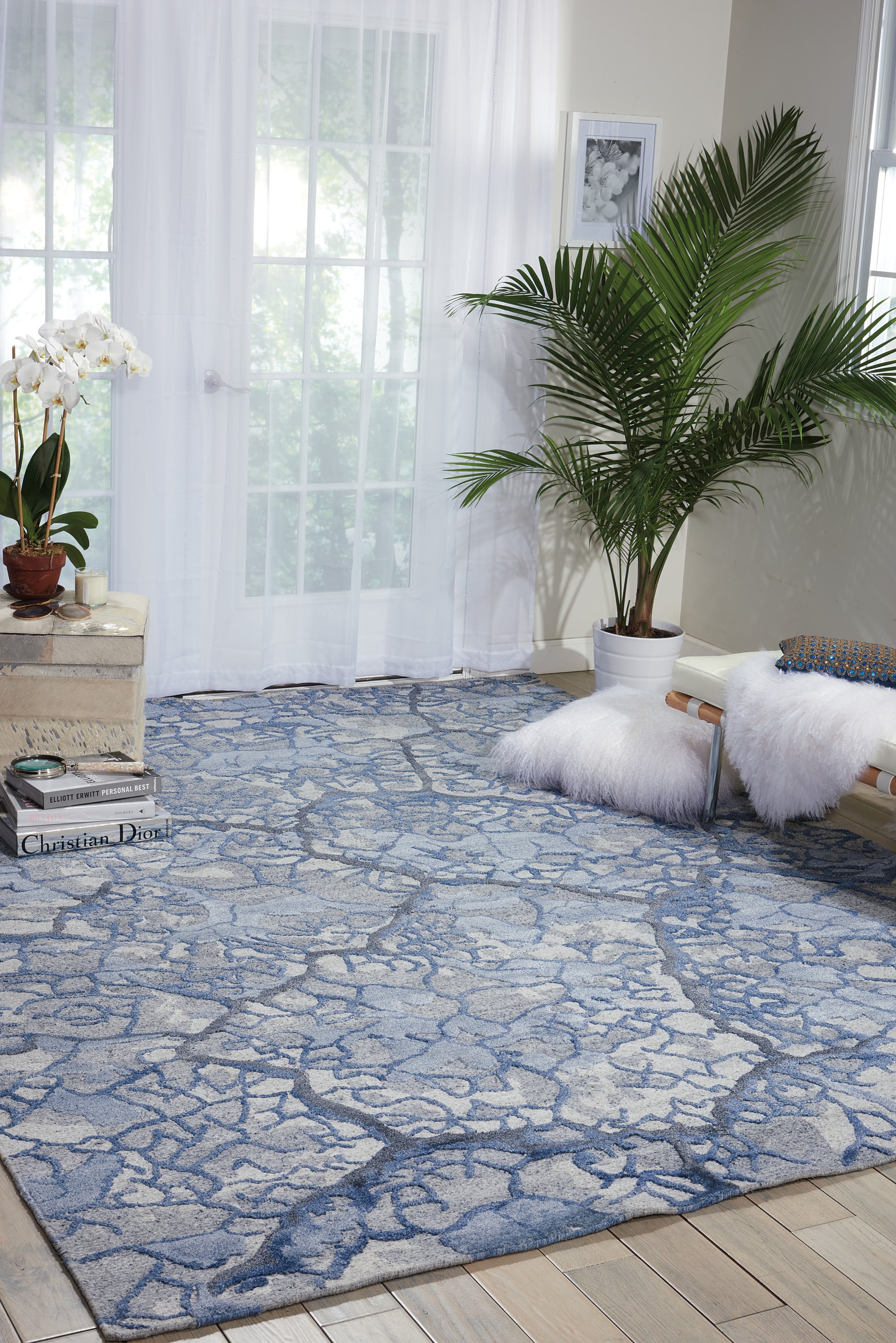Living room with blue gray stone-pattern area rug, white fluffy poufs, potted palm, sheer curtains, and art books.