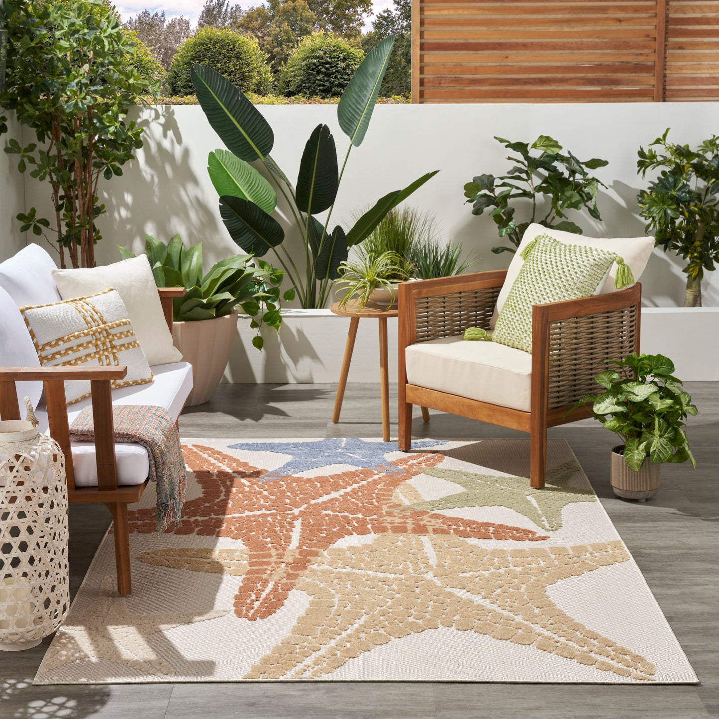 Outdoor patio scene with wicker chairs, white cushions, green pillows, potted plants, and a beige starfish-pattern rug.