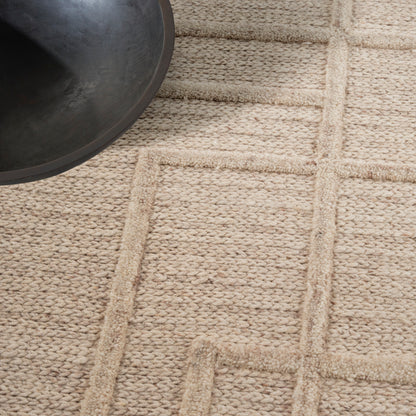 Beige textured jute rug with a geometric grid border; dark metal bowl partially visible in the upper-left.