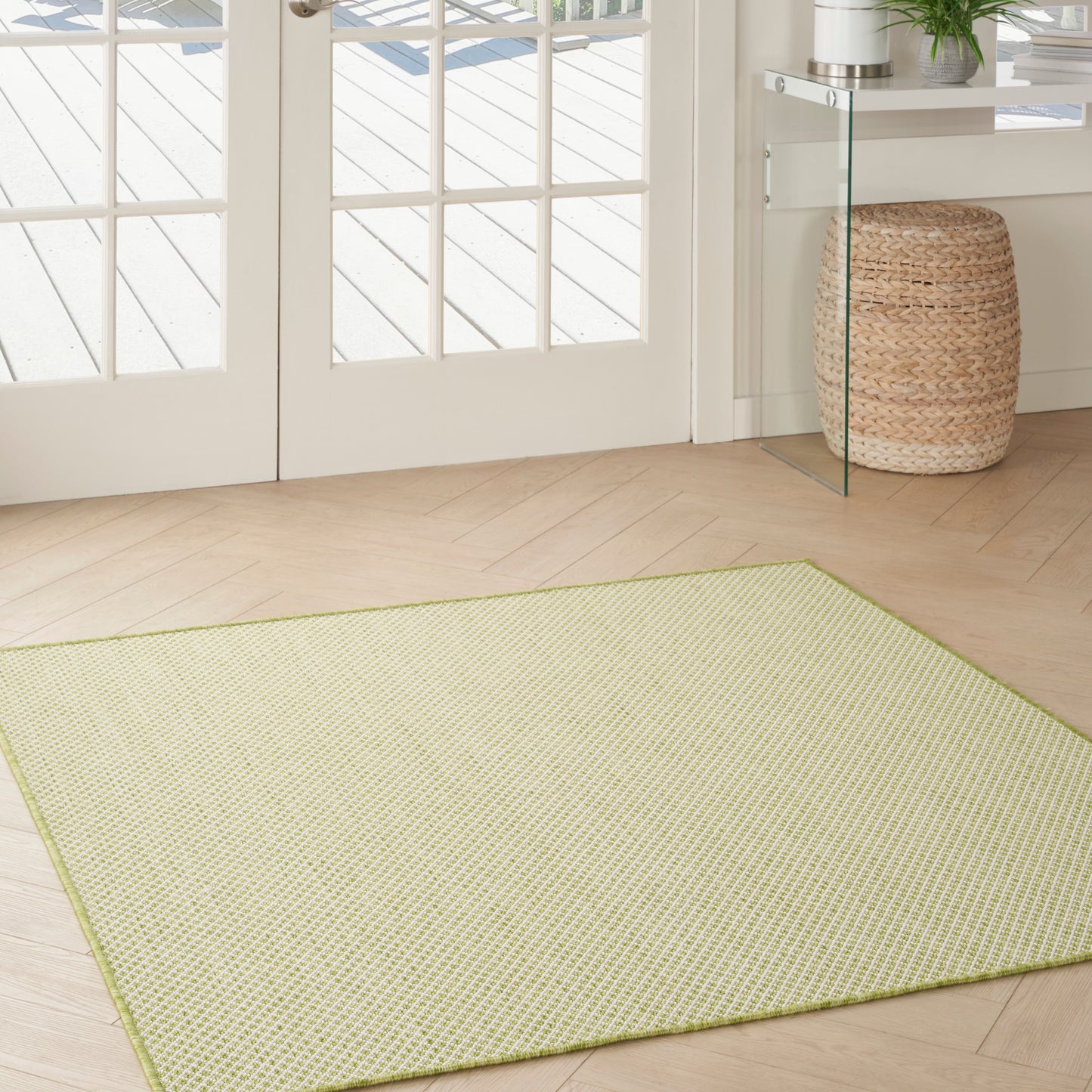 Rectangular light lime woven area rug on pale wood floor beside glass doors, with wicker basket and glass console.