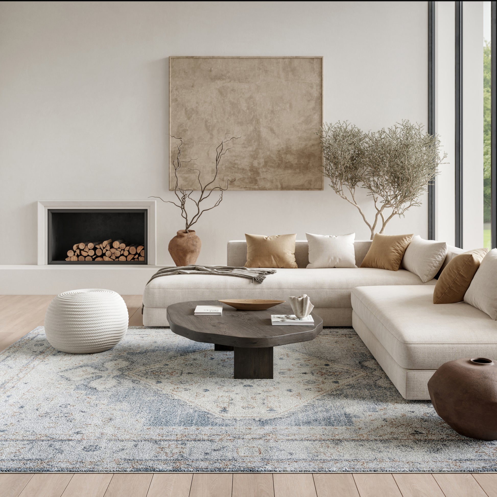 Neutral living room with beige sectional, round dark-wood coffee table, white knitted pouf, blue-gray patterned rug, bare-branch trees in terracotta pots, and tan/cream pillows.