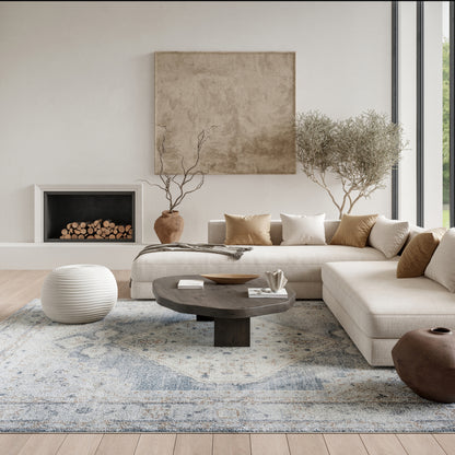Neutral living room with beige sectional, round dark-wood coffee table, white knitted pouf, blue-gray patterned rug, bare-branch trees in terracotta pots, and tan/cream pillows.