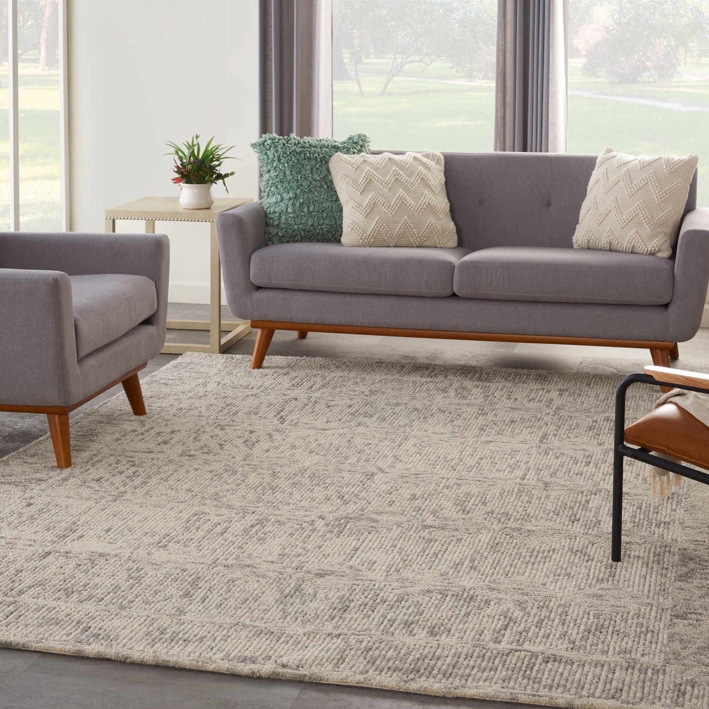 Grey mid-century sofa with matching armchair, two cream zigzag pillows and a green textured pillow, wooden legs, beige rug.