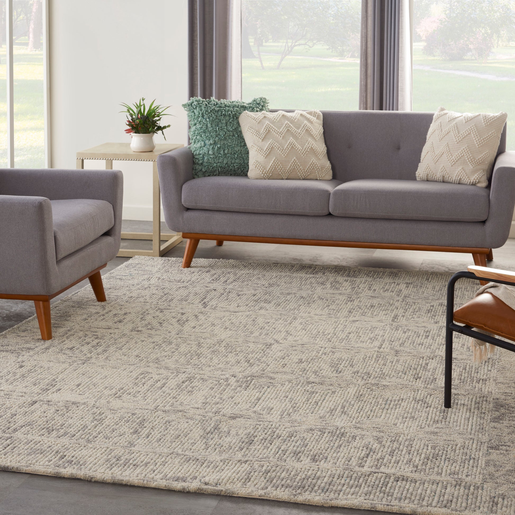 Grey mid-century sofa with matching armchair, two cream zigzag pillows and a green textured pillow, wooden legs, beige rug.