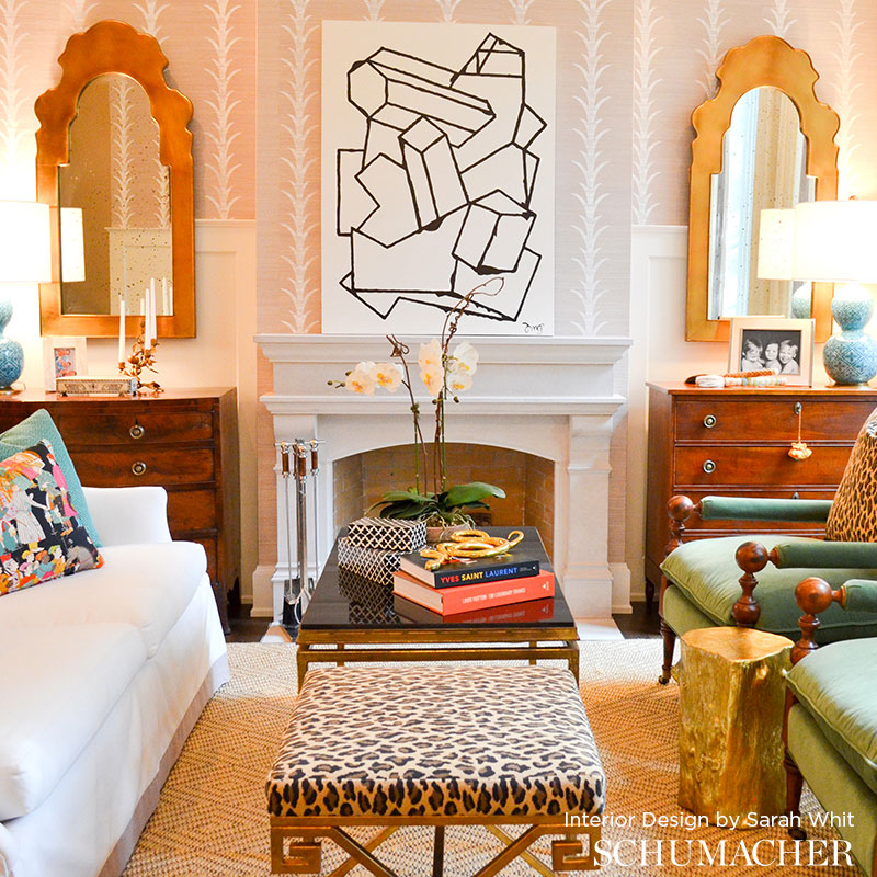 Eclectic living room with leopard-print bench, glass coffee table, pink wallpaper, white fireplace, gold-framed mirrors.