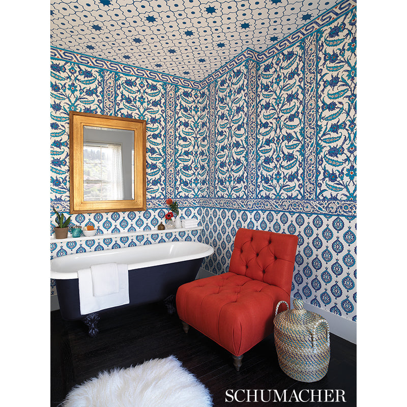 Blue and white patterned wallpaper surrounds a navy clawfoot tub, red tufted chair, gold mirror, woven basket, and white rug.