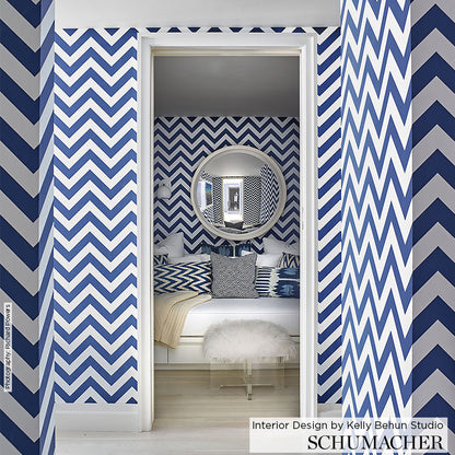 Blue and white chevron wallpaper across walls; round wall mirror, white bed with blue pillows, fluffy white ottoman.