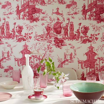 Pink-red toile wallpaper featuring pagodas and bridges; white vase, pink glassware, teacup, and green bowl on a dining table.