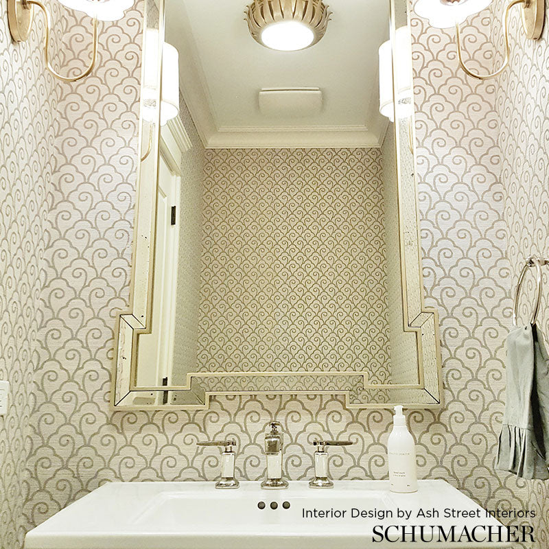 Beige patterned wallpaper in a vintage bathroom with a large beveled-framed mirror above a white sink & chrome faucets.