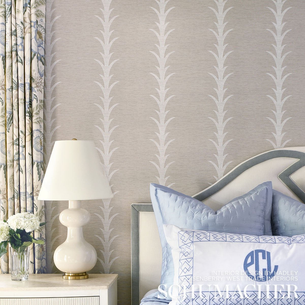 Bedroom with pale gray wallpaper pattern, curved gray-trim headboard, blue pillows, white lamp on a nightstand, floral curtains.